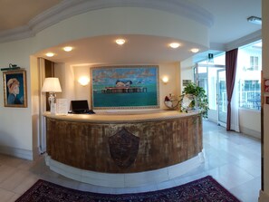 A reception desk with a painting of a pier, a lamp, and a plant. - Hotel Greif (Lignano Sabbiadoro)