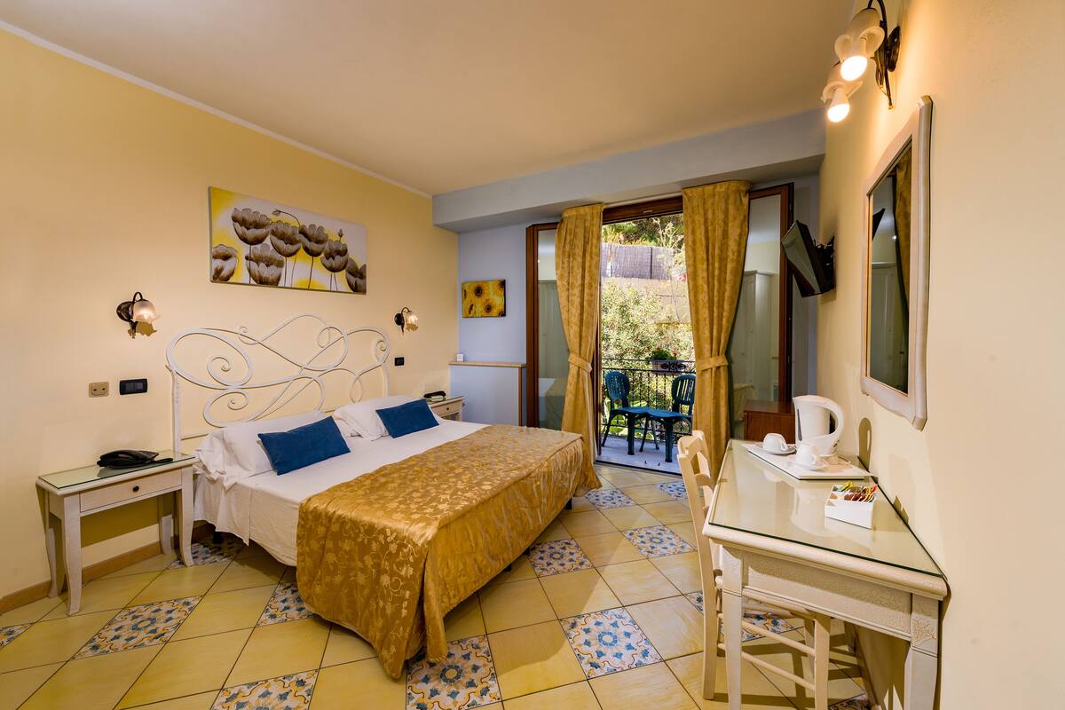 Ein Hotelzimmer mit Bett, Nachttisch und Schreibtisch mit Stuhl. Es gibt einen Balkon mit Stuhl und Tisch, sowie ein Bild an der Wand.