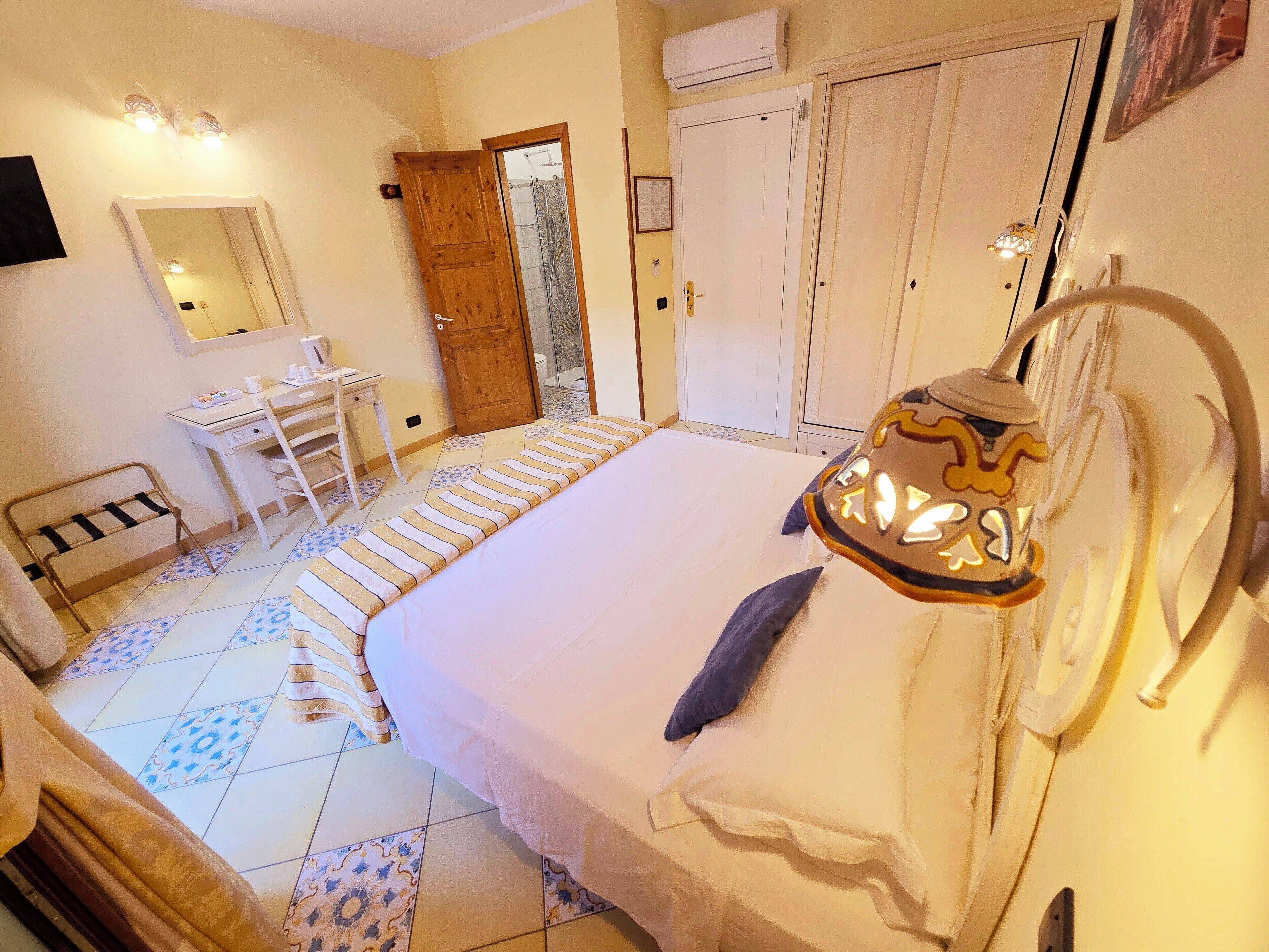 A bedroom with a bed, bedside table, and a mirror. There is a bathroom visible through an open door.