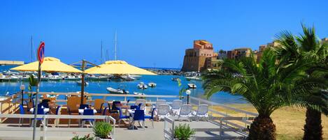 A marina with boats, a café with outdoor seating, and a historic building in the background.
