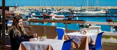 Comedor con mesas y sillas, con vista a un puerto deportivo y barcos.