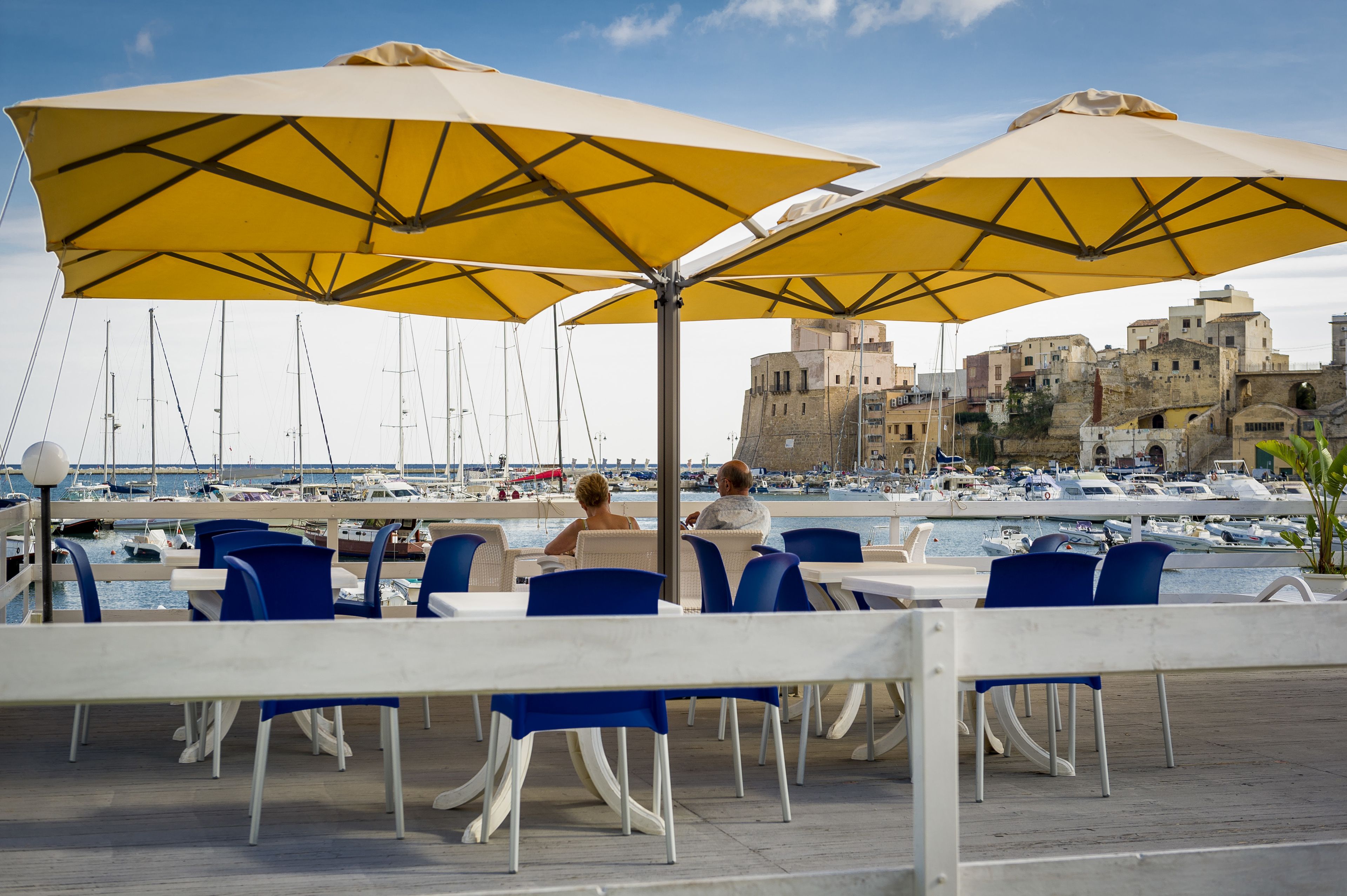 Un espace de restauration en bord de mer, avec des tables et des chaises sous des parasols jaunes, offrant une vue sur un port de plaisance avec des bateaux et des bâtiments historiques.