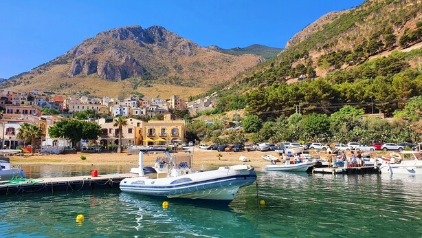Une ville côtière où des bateaux sont amarrés dans un port de plaisance, avec un hôtel imposant et un paysage montagneux en arrière-plan.