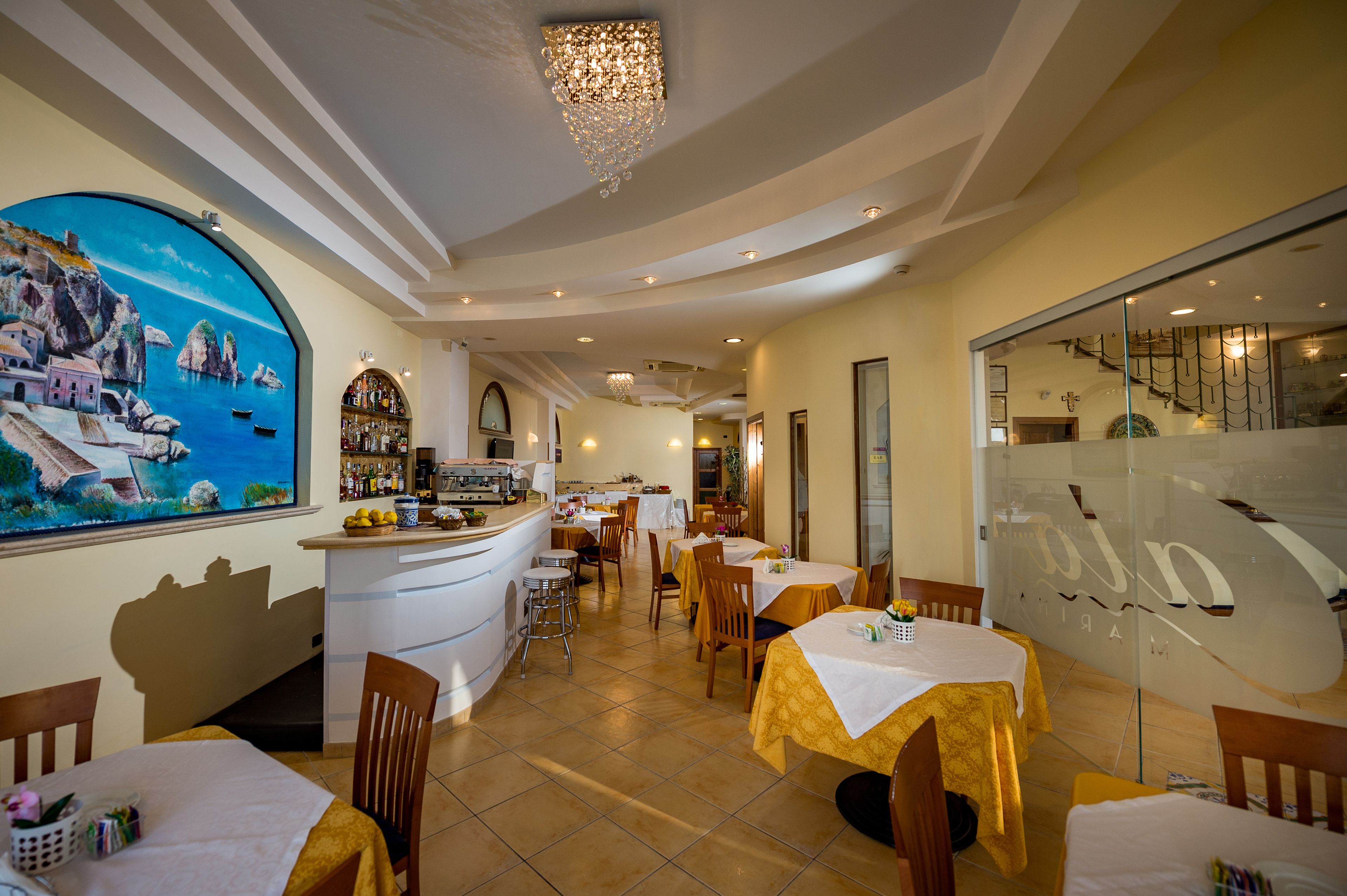 Un restaurante con un mural náutico, una zona de bar y varias mesas de comedor con manteles amarillos.