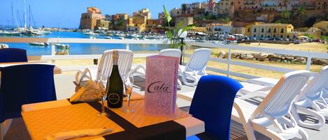 A table set with a bottle of wine, glasses, a menu, and a hotel brochure, overlooking a marina with boats and coastal buildings.