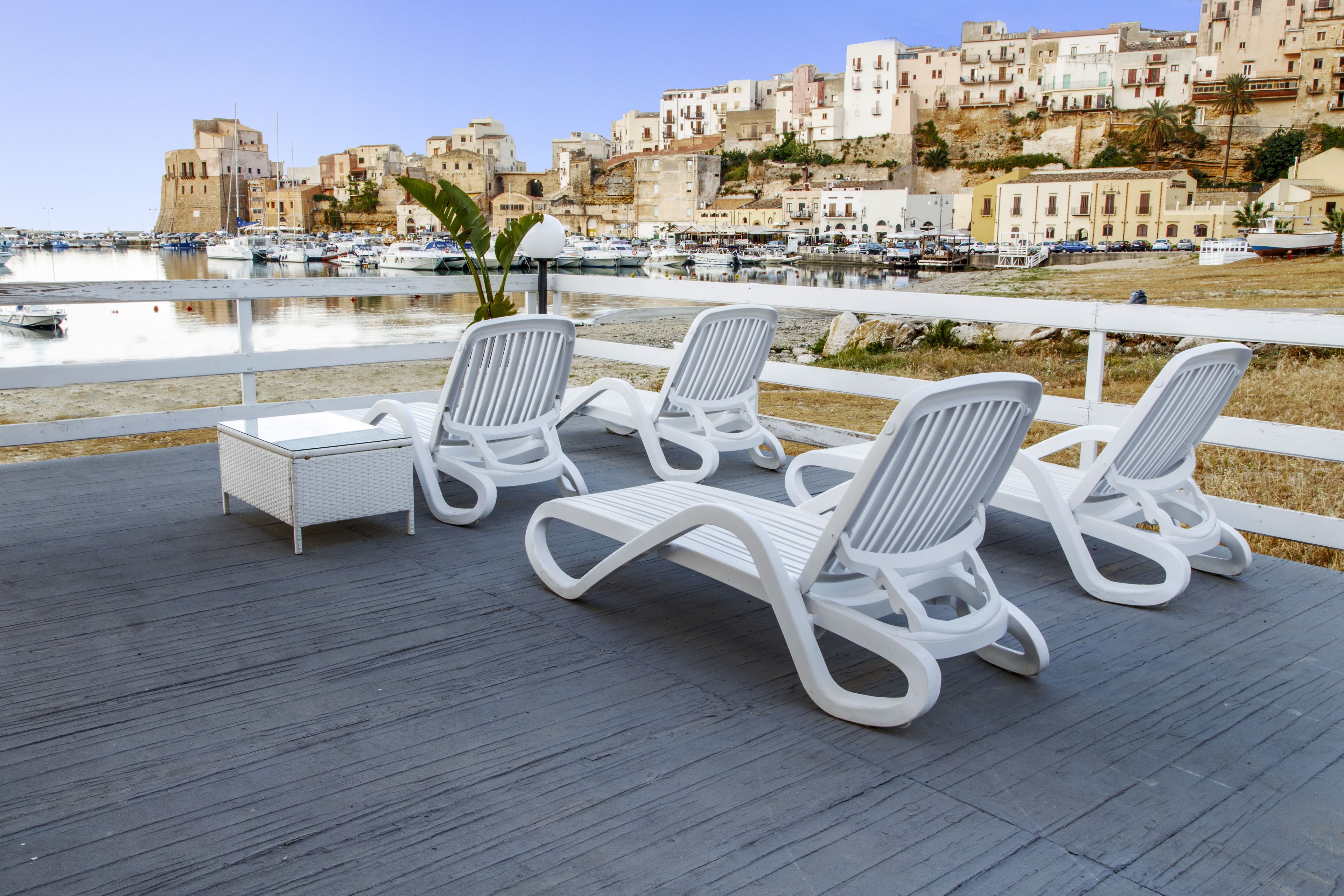 Una terraza con sillones blancos y una pequeña mesa, con vista a un puerto deportivo con barcos y edificios al fondo.