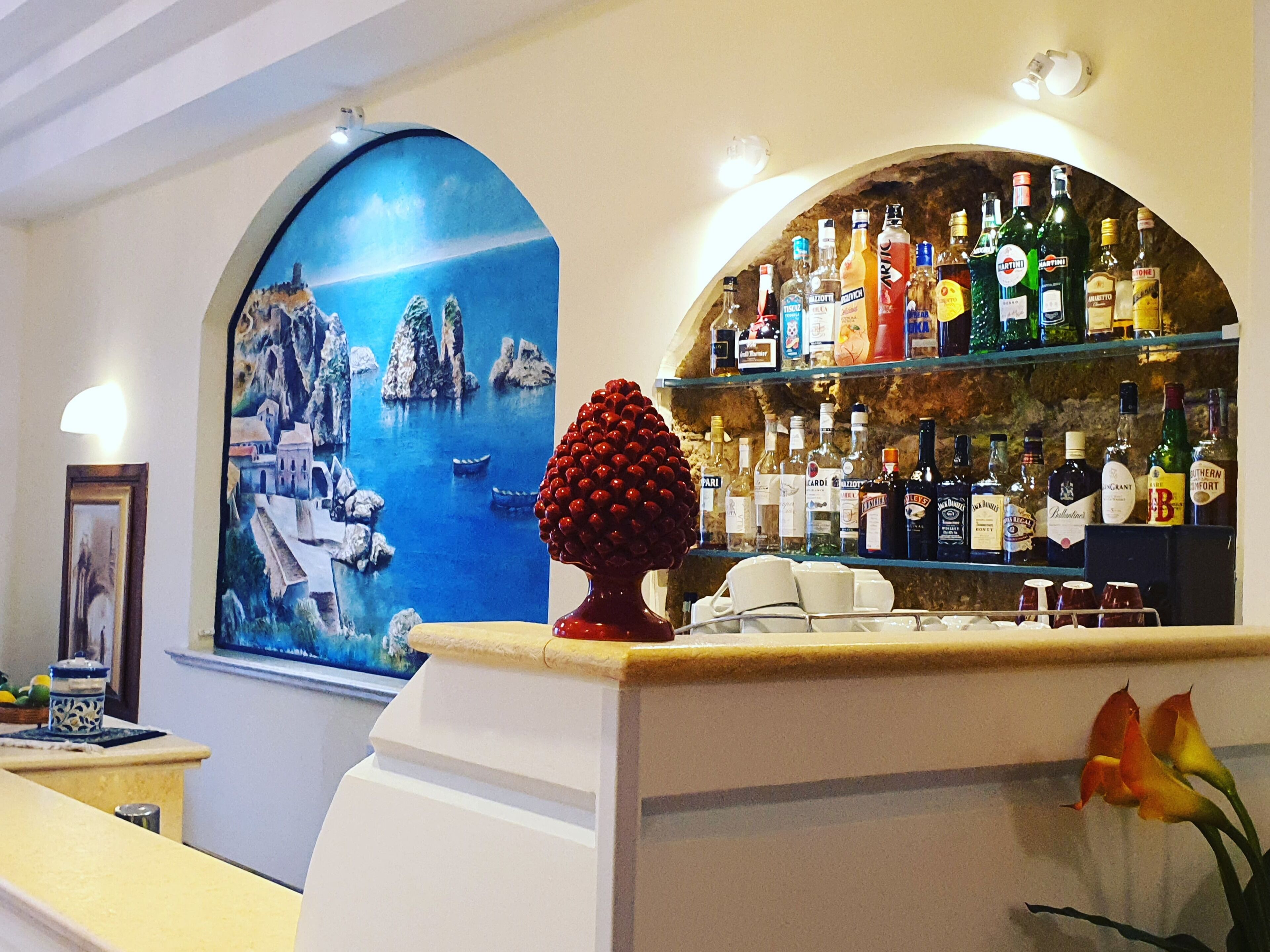 Uma área de bar com um mural de paisagem costeira, variedade de garrafas de bebidas alcoólicas e um objeto decorativo vermelho no balcão.