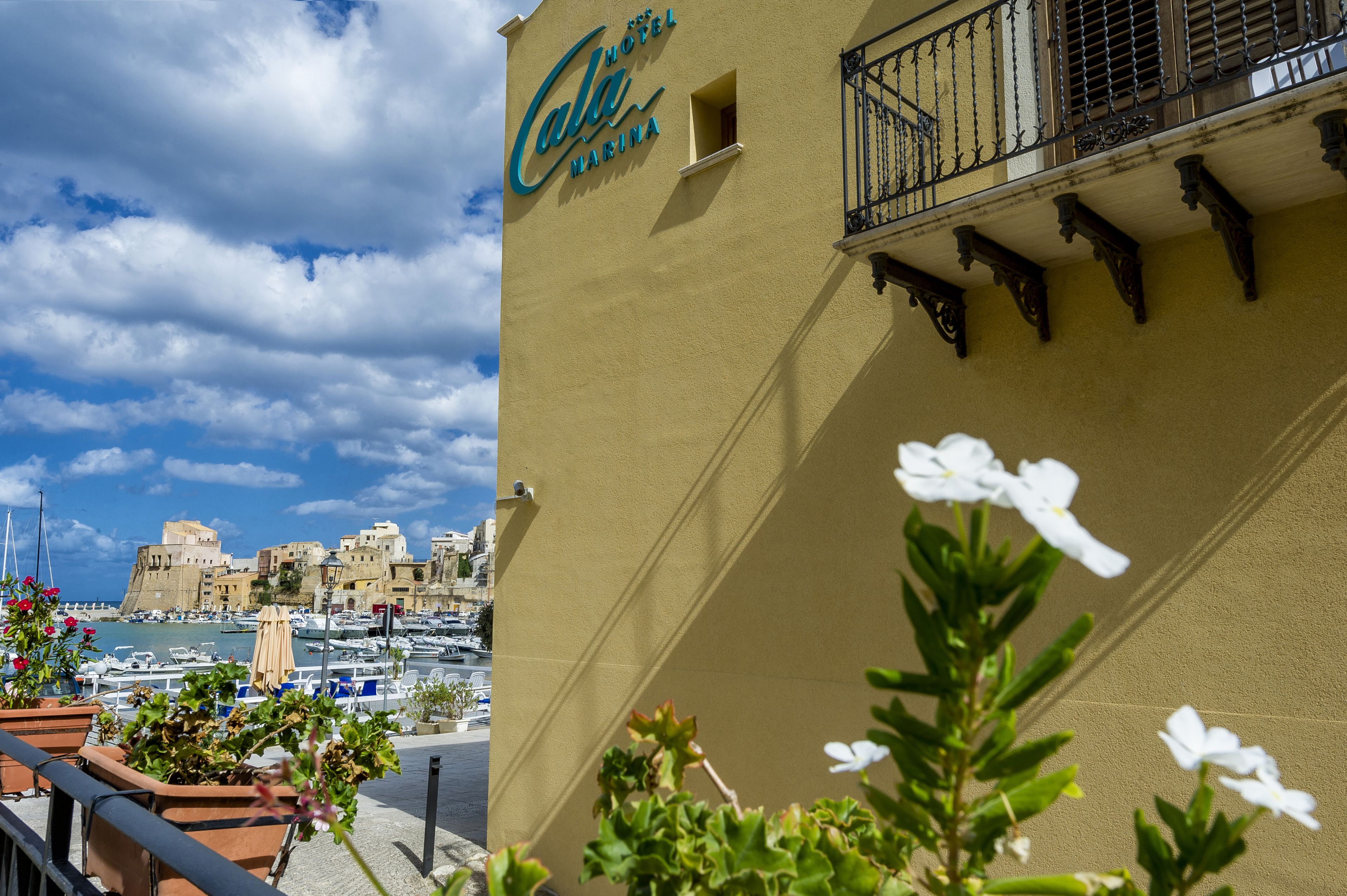 A hotel named "Hotel Cala Marina" with a balcony overlooking a marina and historic buildings.