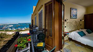 Un balcon avec vue sur la mer, un lit avec du linge de maison blanc et des coussins turquoise, et un tableau accroché au mur.