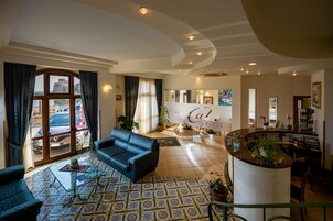 Ein geräumiger Wohnbereich mit einem blauen Sofa, einem Couchtisch und einem gemusterten Boden. Ein großes Fenster mit Vorhängen ist zu sehen, und im Hintergrund befindet sich ein Empfangstresen mit dem Schriftzug "Café".