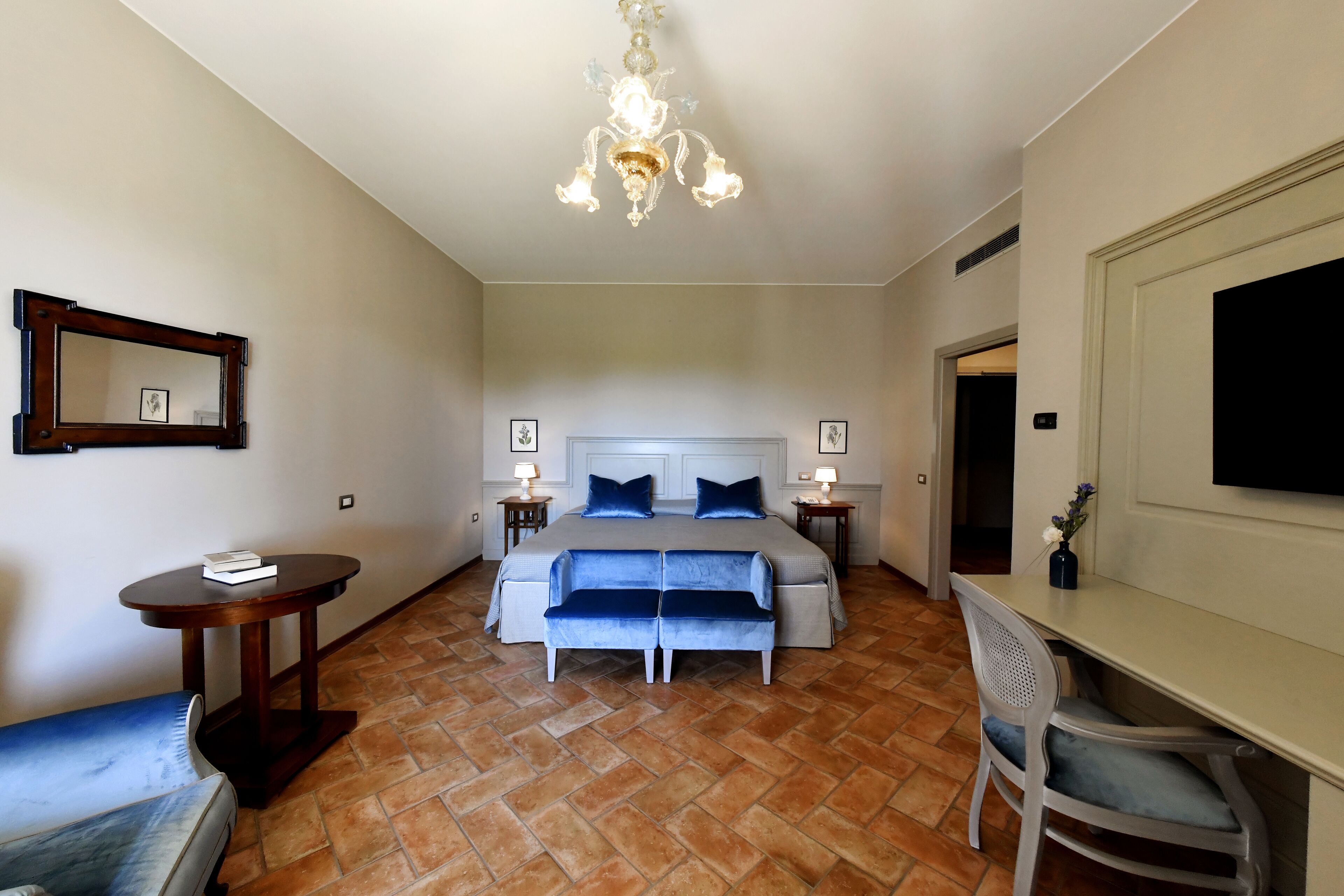 A hotel room with a large bed, a desk, a chair, a small table, a mirror, and a chandelier.