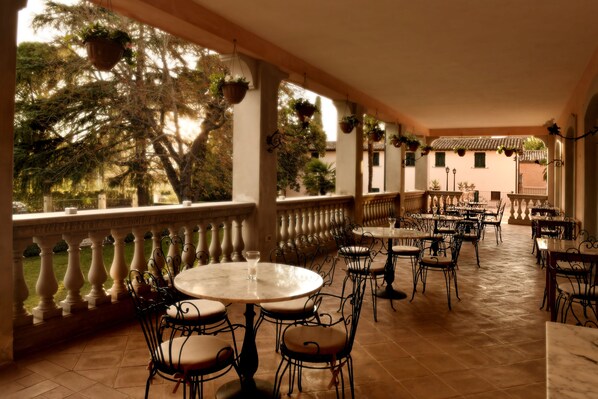 An outdoor patio with round tables and chairs, surrounded by a balustrade, and potted plants hanging from the ceiling.