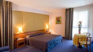A hotel room with a bed, bedside tables, a small table with a chair, and a window with curtains.