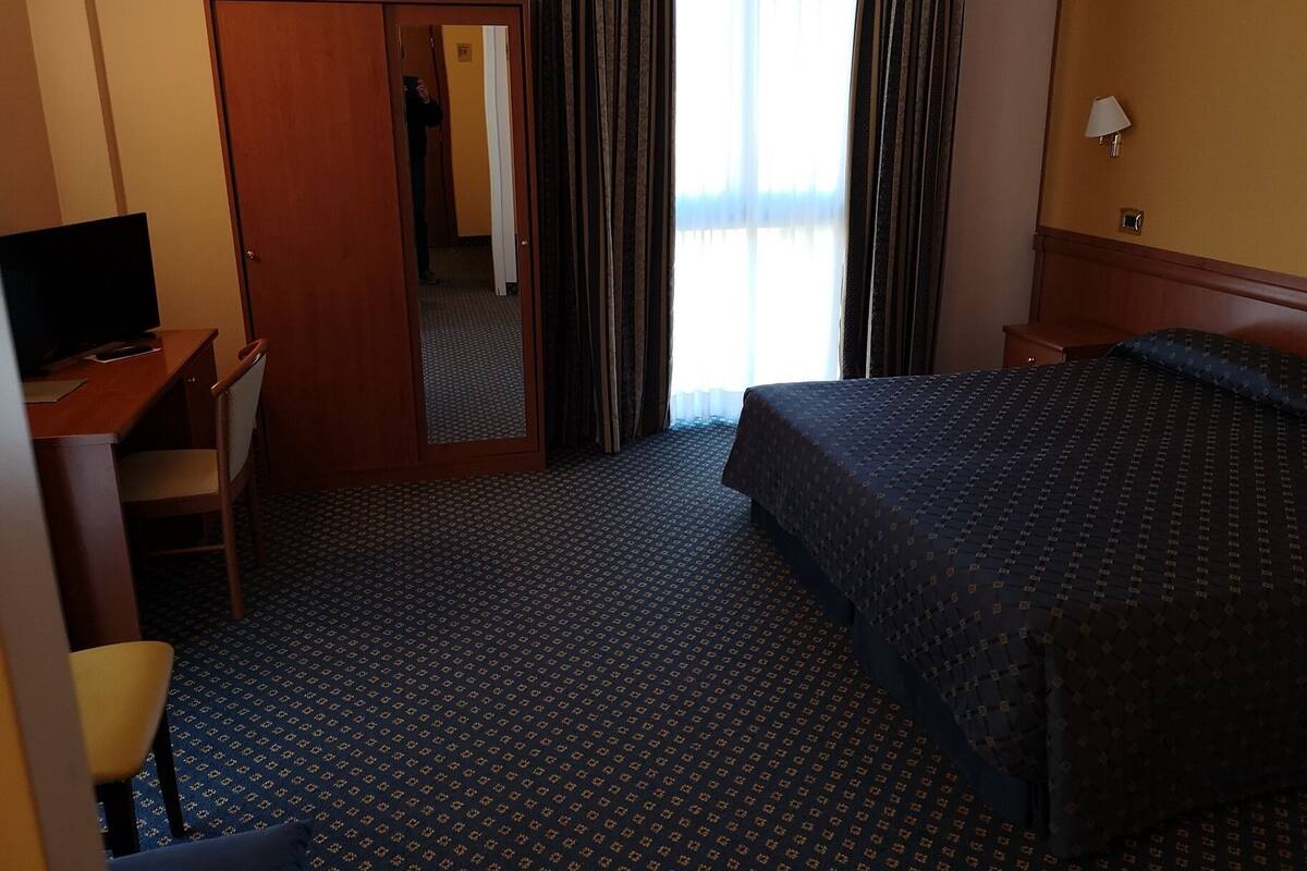 A hotel room with a bed, a desk with a computer, a chair, a wardrobe, and a mirror.