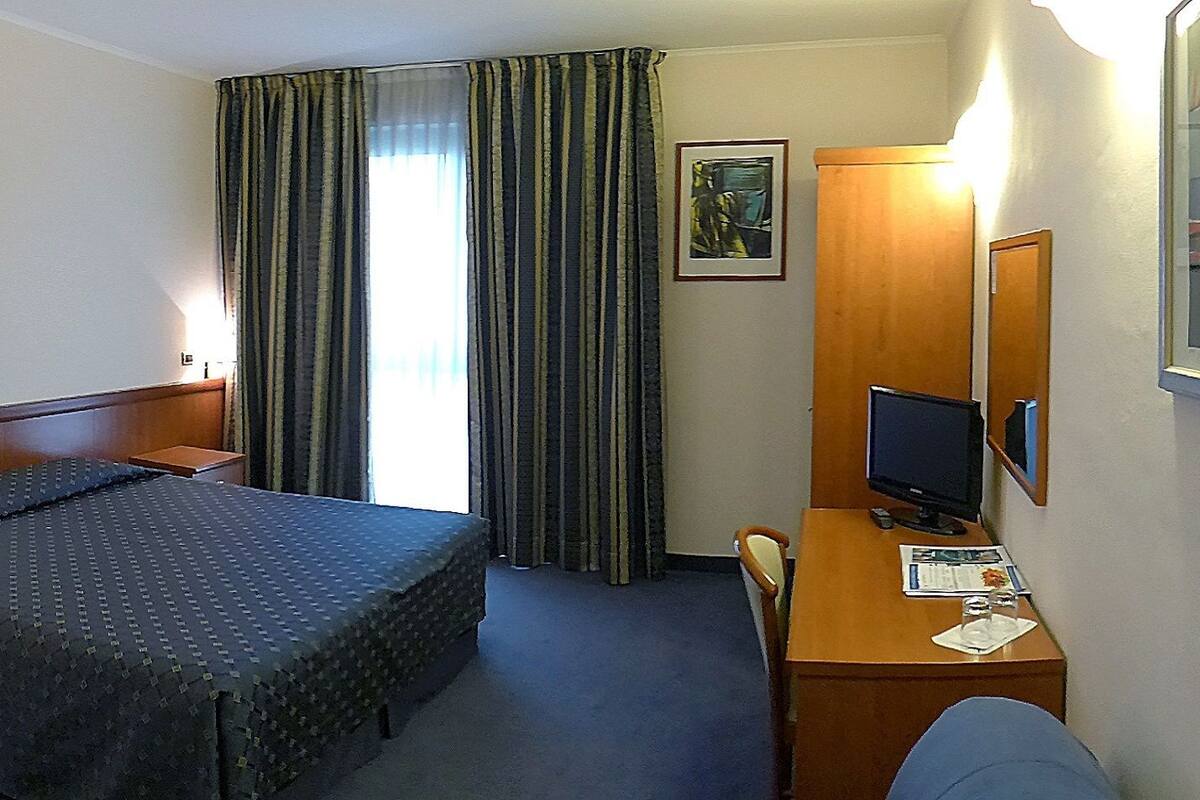 A hotel room with a bed, a desk with a computer, a chair, a television, and a window with curtains.