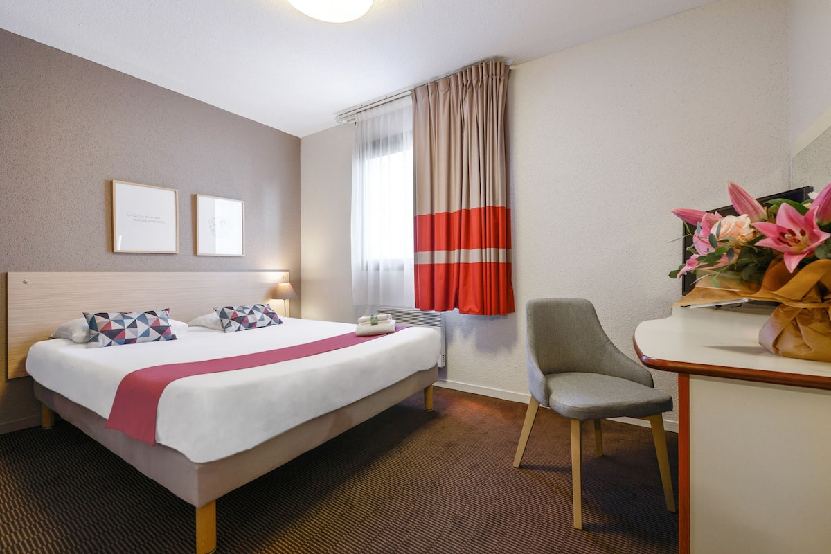 Habitación de hotel con cama, escritorio, silla, ventana con cortinas y pared decorada con cuadros.