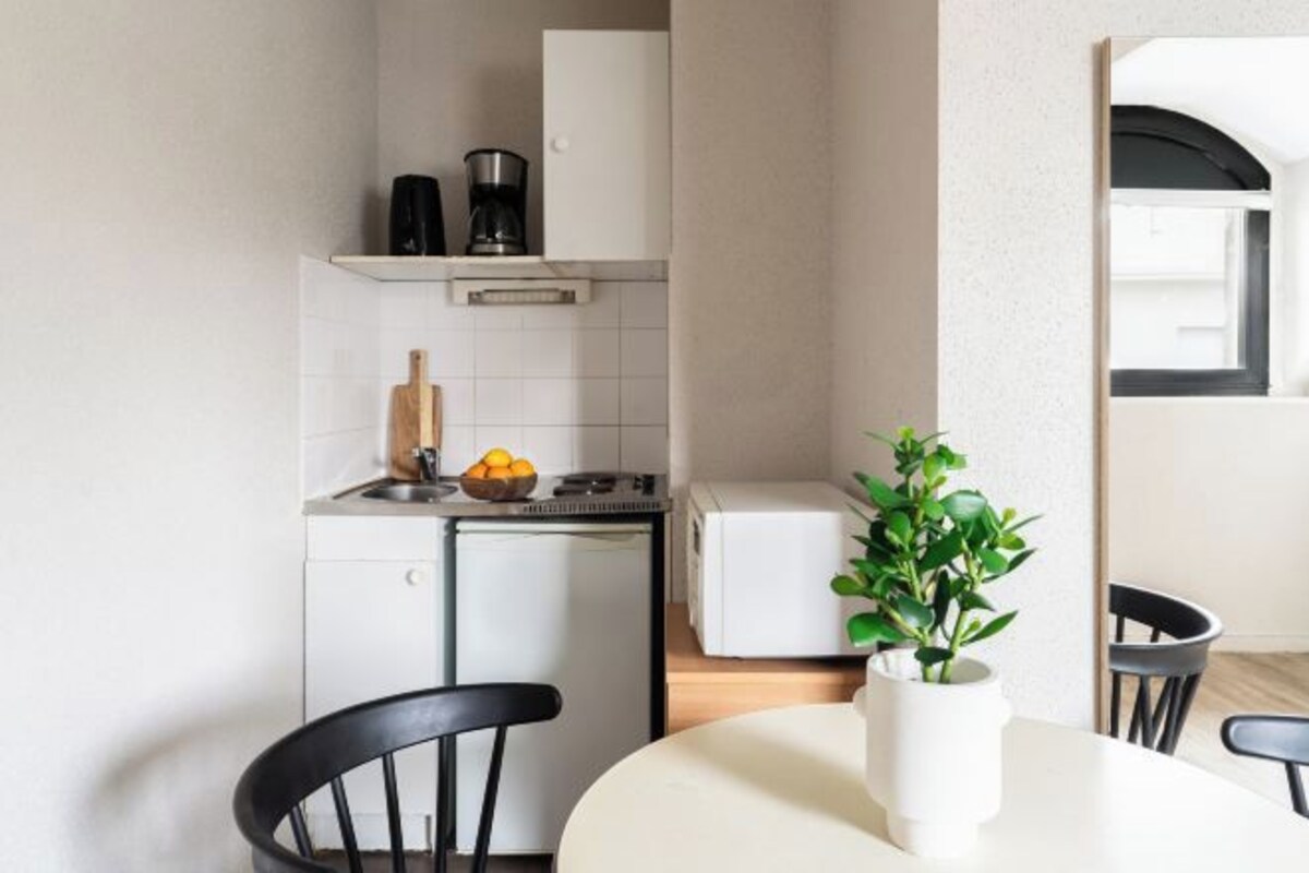 Una cocina compacta con un refrigerador blanco, una cafetera y una estufa pequeña. Hay una mesa de comedor con sillas y una planta en maceta sobre la mesa.
