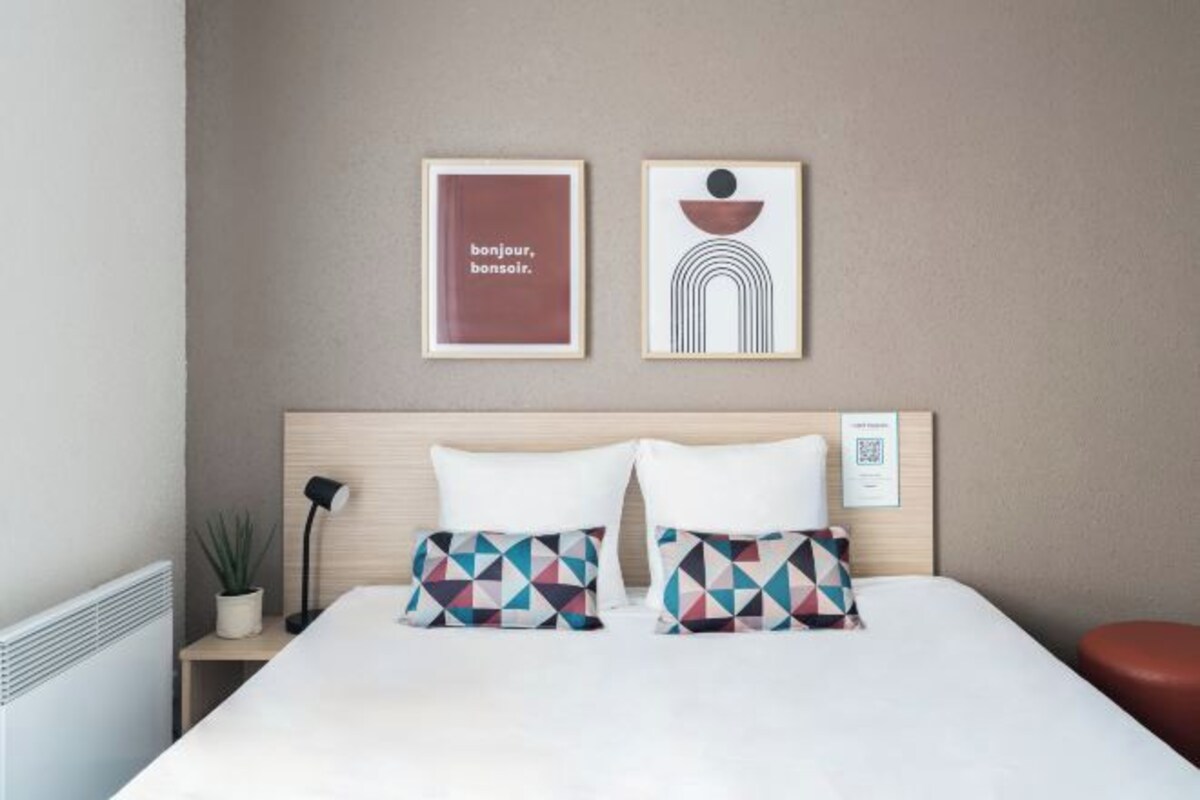 Habitación de hotel con una cama, dos almohadas, una mesita de noche con una lámpara y dos cuadros enmarcados en la pared.