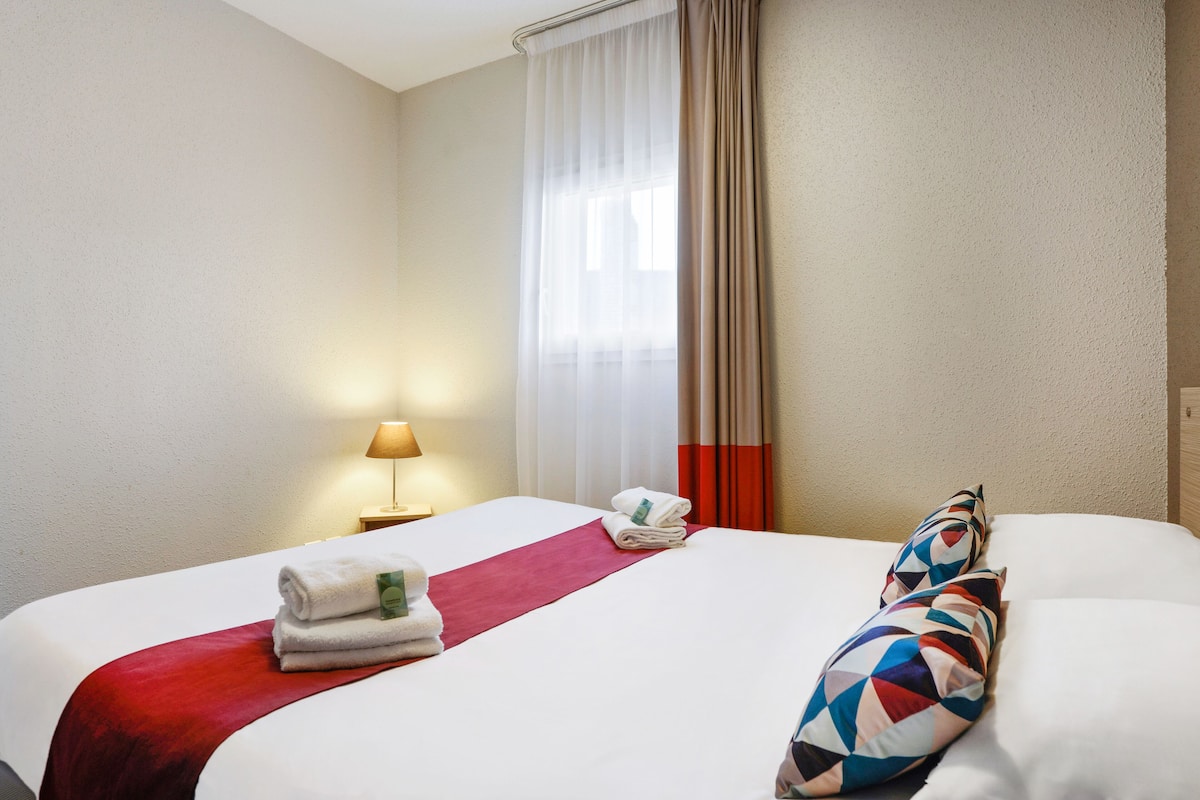 Una habitación de hotel con una cama bien hecha, una manta roja y un cojín estampado.