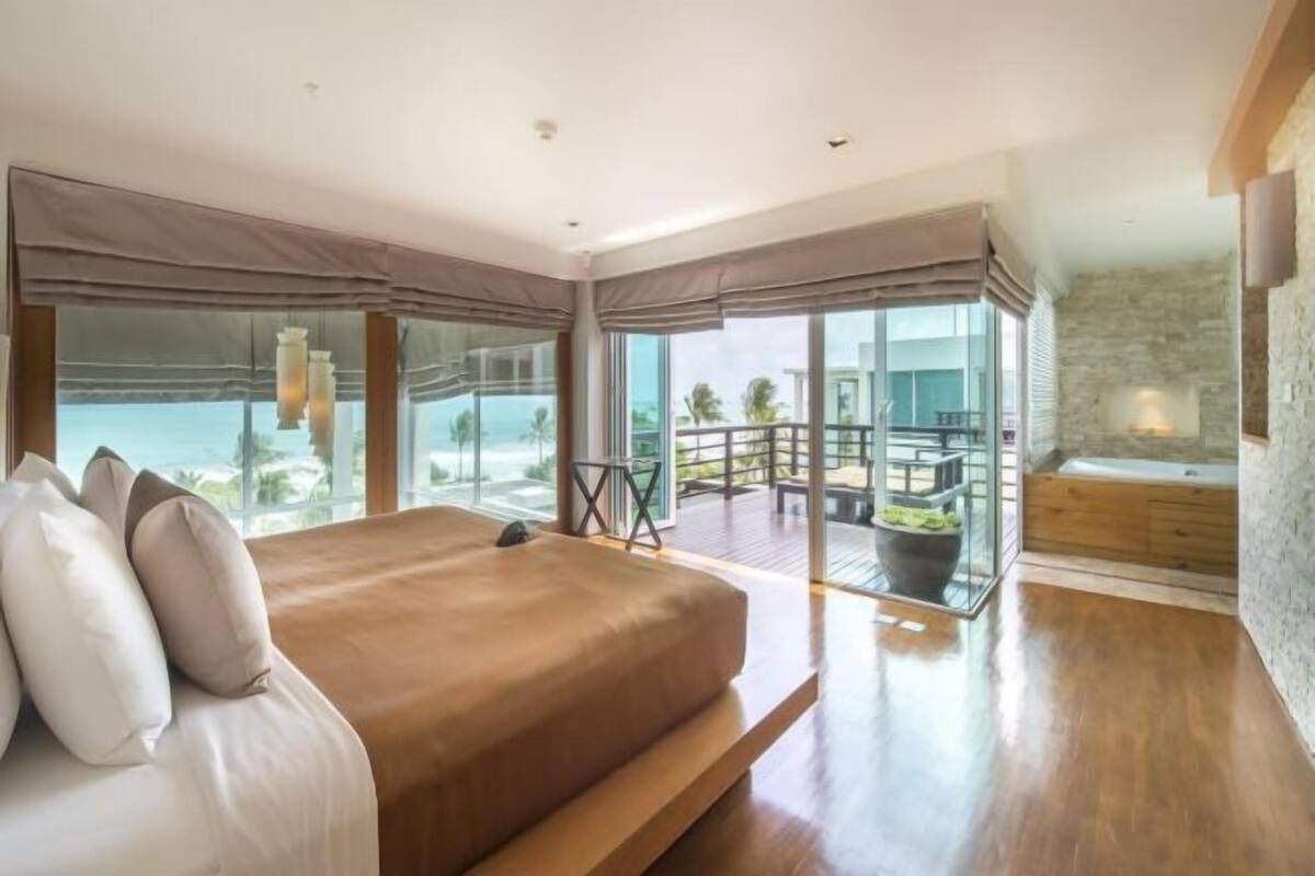 A bedroom with a large bed, a balcony with a view of the ocean, and a stone wall.