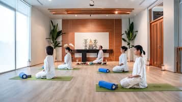 A yoga studio with participants seated on mats, a wooden wall with a Buddha statue, and large windows.