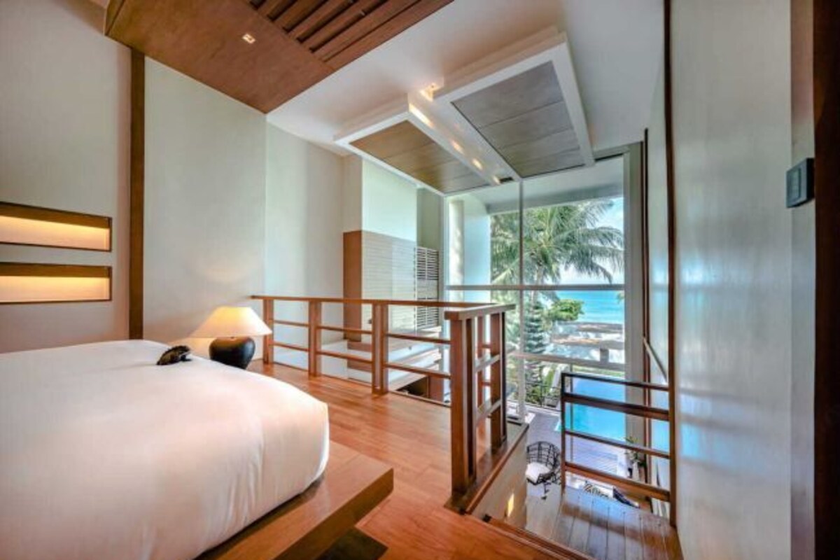A hotel room with a bed, a nightstand, a lamp, a balcony with a view of the ocean and palm trees.