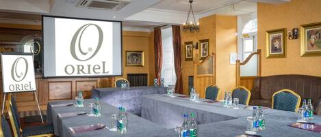 Un salón de conferencias con una larga mesa, sillas, botellas de agua y una pantalla proyectora mostrando el logotipo "ORIEL".