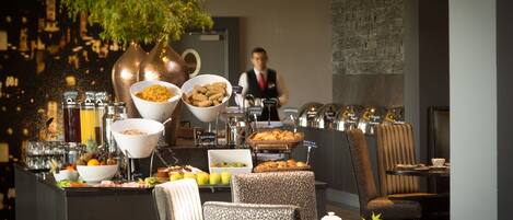 A buffet setup with various food items, seating arrangements, and a waiter in the background.