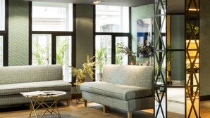 Una sala de estar moderna con sofá, sillón y mesa de centro. Hay ventanas amplias con plantas y un panel decorativo en la pared.