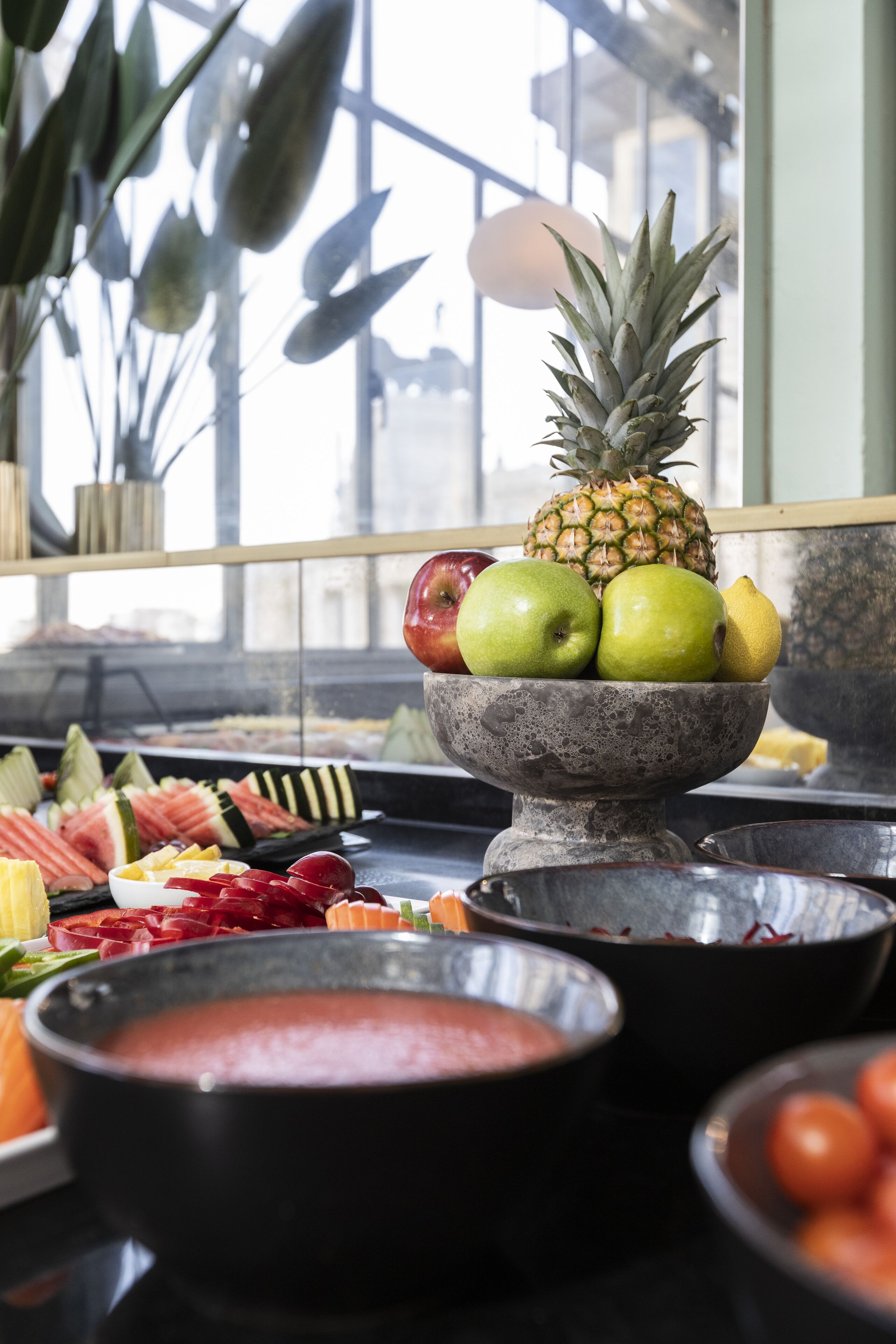 A fruit bowl with apples, a pineapple, and a pear on a table with various fruits and a bowl of soup.