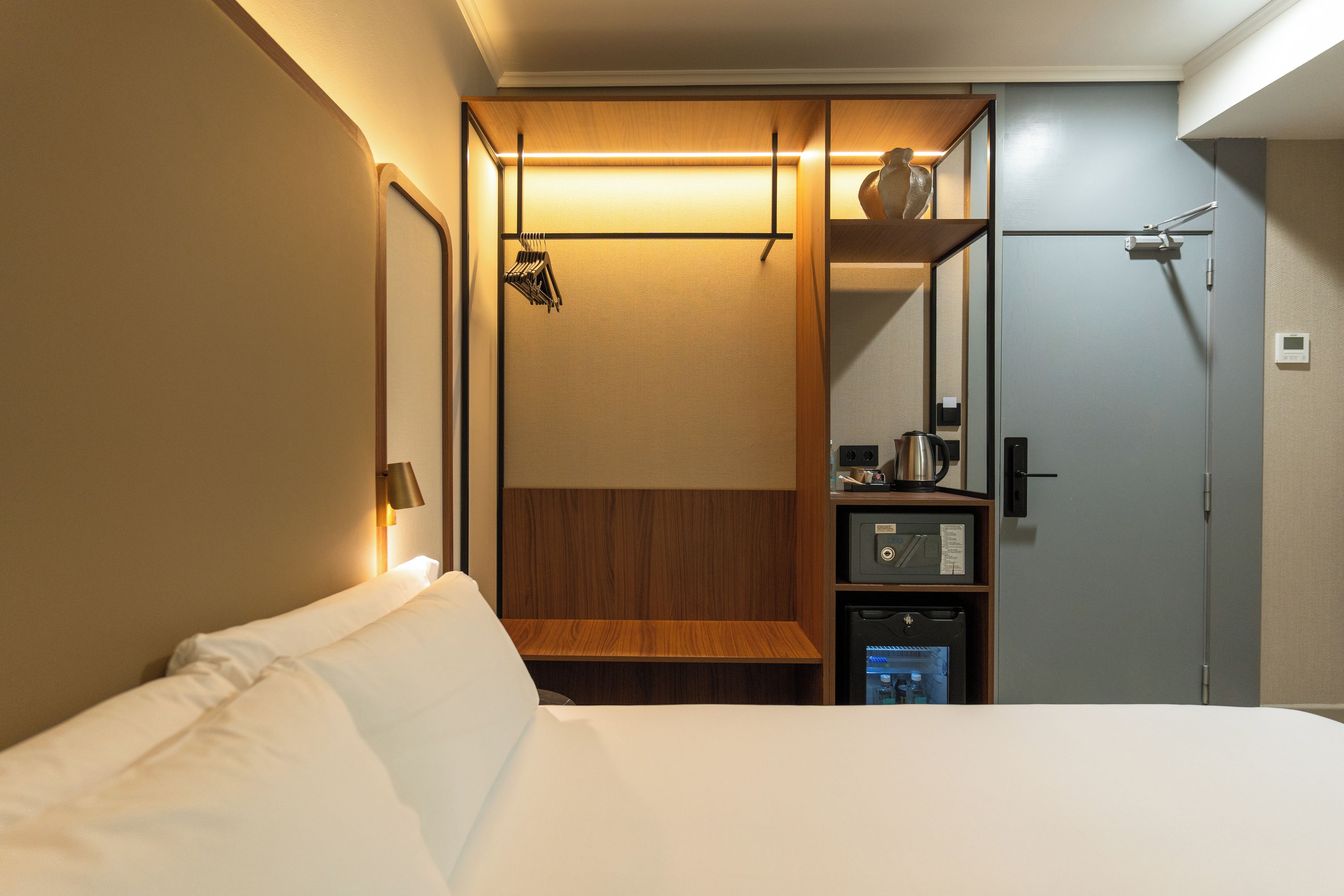 Une chambre d’hôtel avec un lit, une étagère en bois, une bouilloire, un petit réfrigérateur et un miroir.