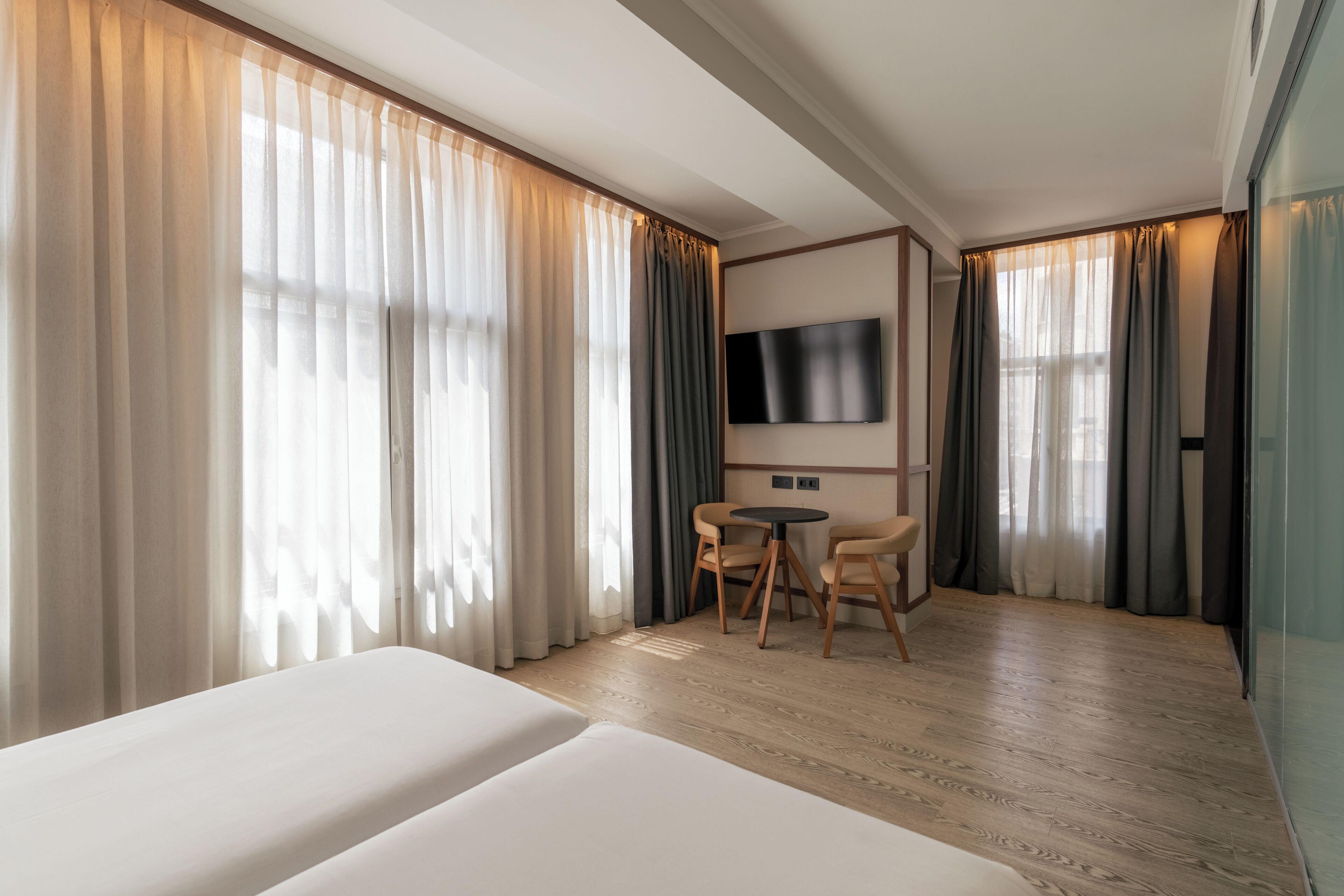 Habitación de hotel con dos camas, un televisor montado en la pared, una mesita redonda con sillas y ventanas amplias con cortinas.