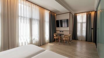 Habitación de hotel con dos camas, un televisor montado en la pared, una mesita redonda con sillas y ventanas amplias con cortinas.