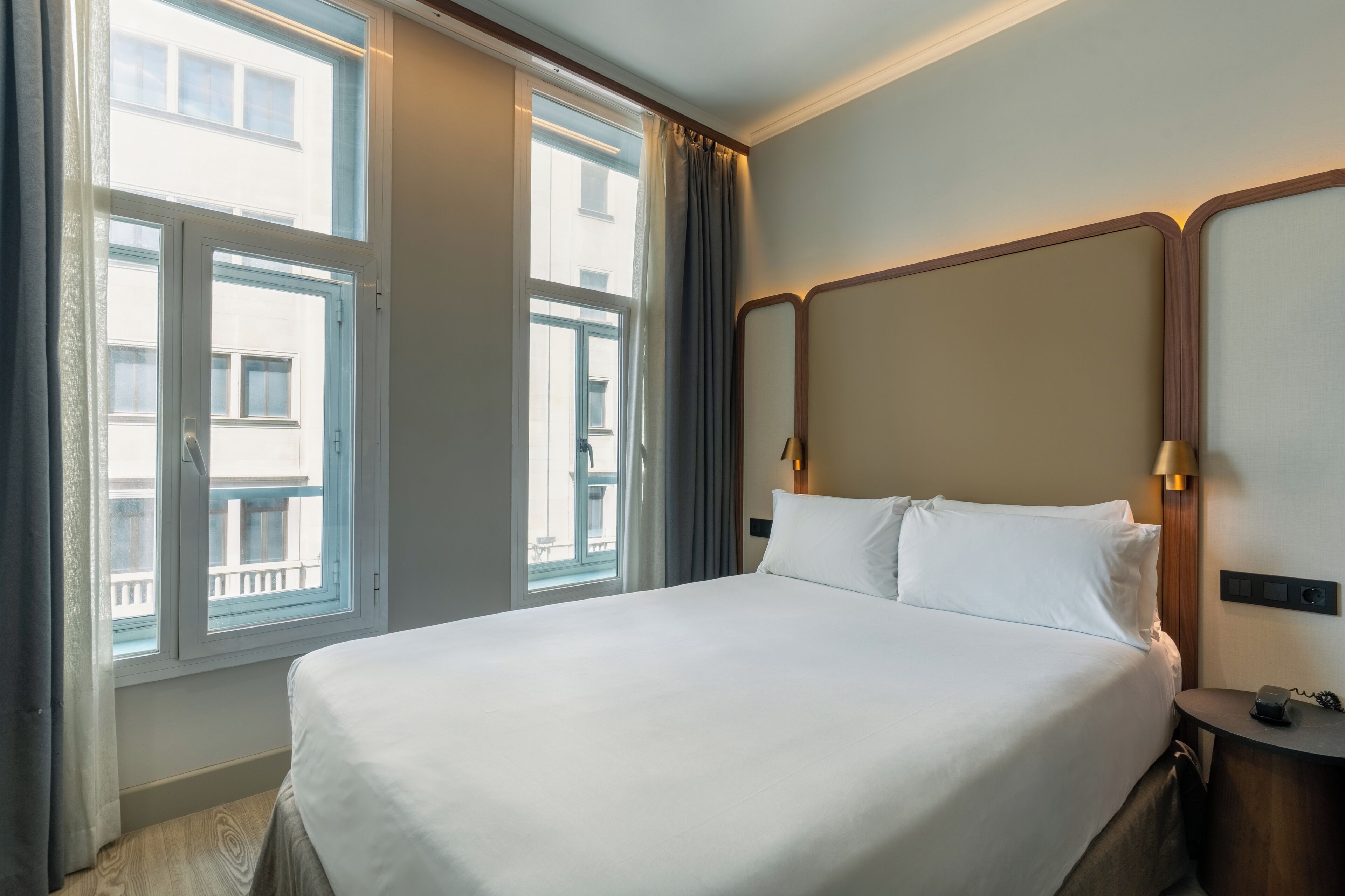Una habitación de hotel con una cama grande, un cabecero de madera, una mesita de noche con lámpara y una ventana con cortinas.