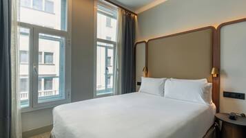 Una habitación de hotel con una cama grande, un cabecero de madera, una mesita de noche con lámpara y una ventana con cortinas.