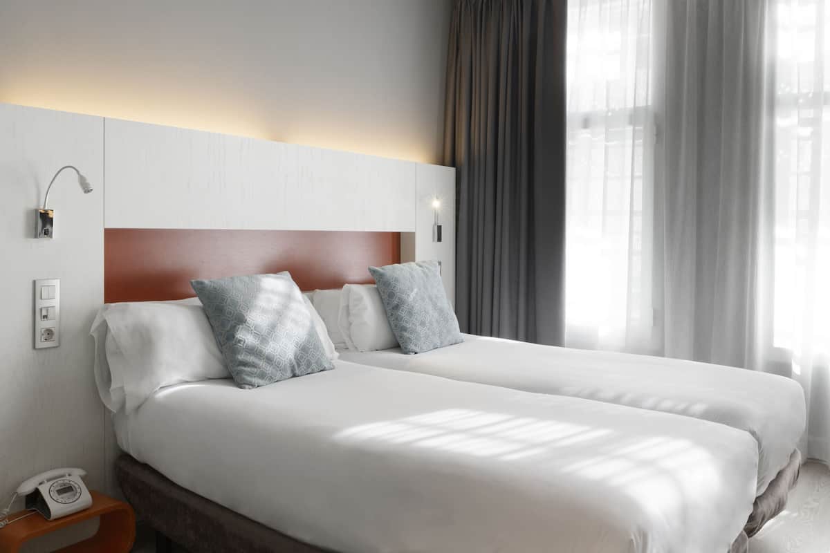 Habitación de hotel con dos camas, cabecero de madera, mesita de noche con teléfono y ventana con cortinas.