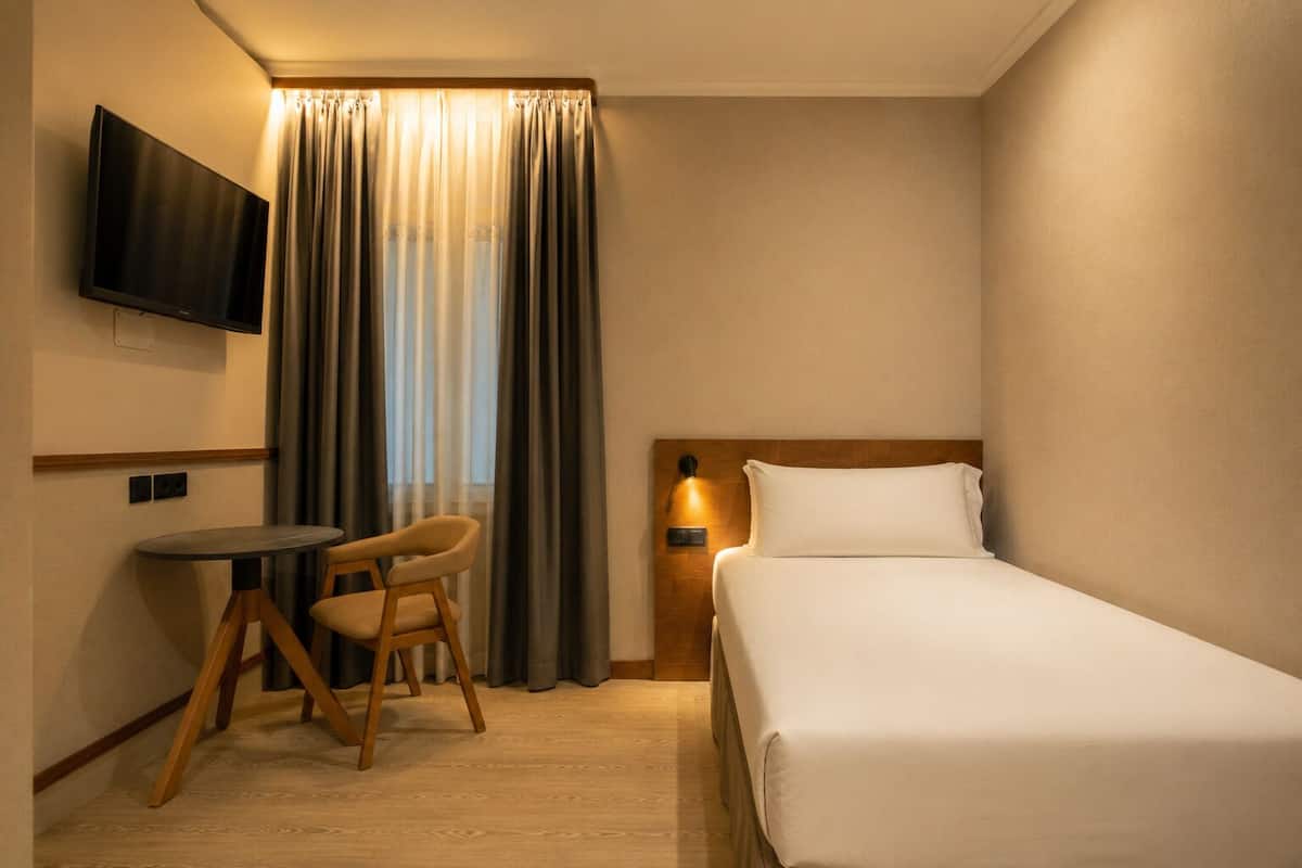 Habitación de hotel con cama, escritorio con silla, televisor montado en la pared y ventana con cortinas.
