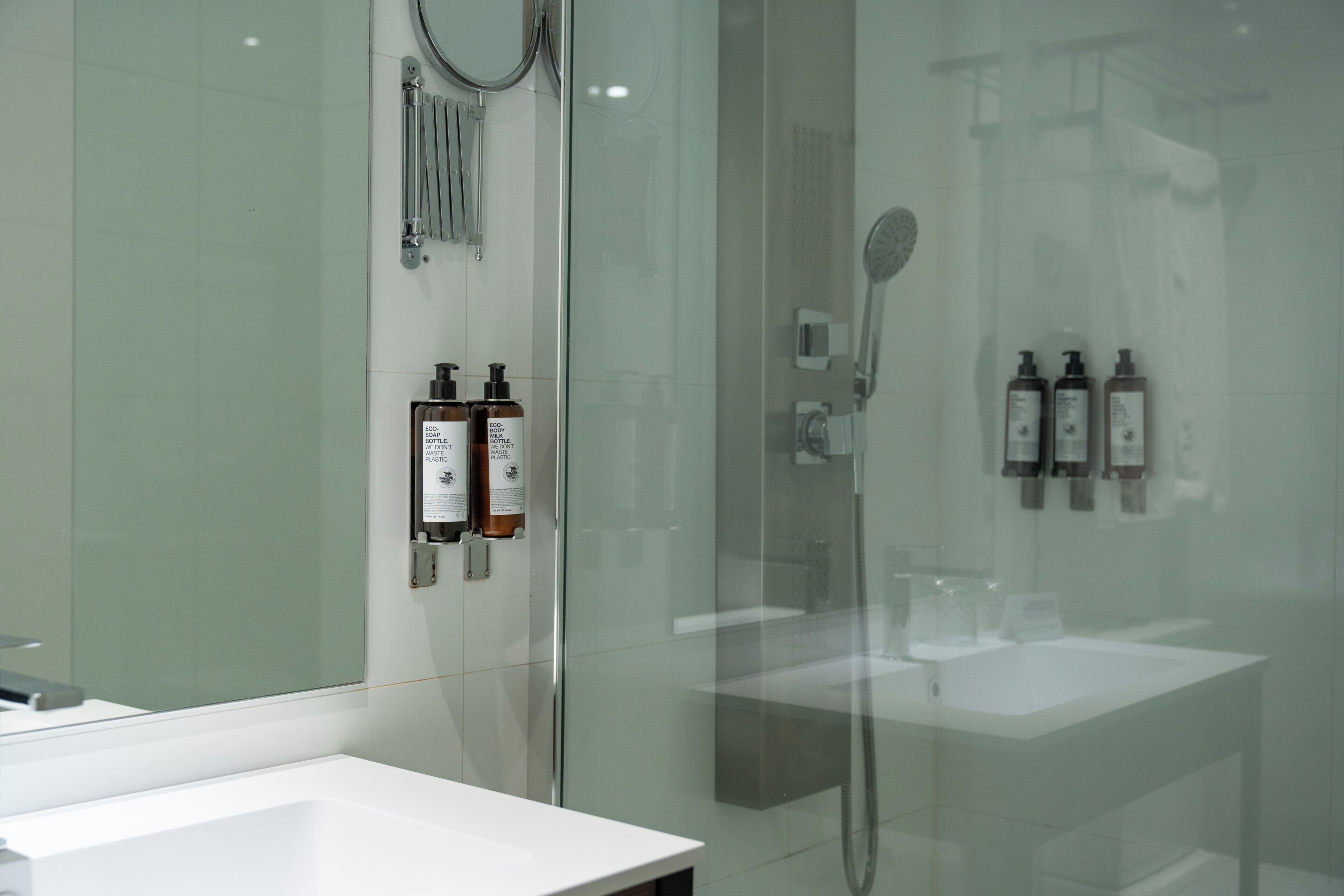 Un bagno moderno con doccia vetrata, lavandino bianco e due flaconi di sapone ecologico.