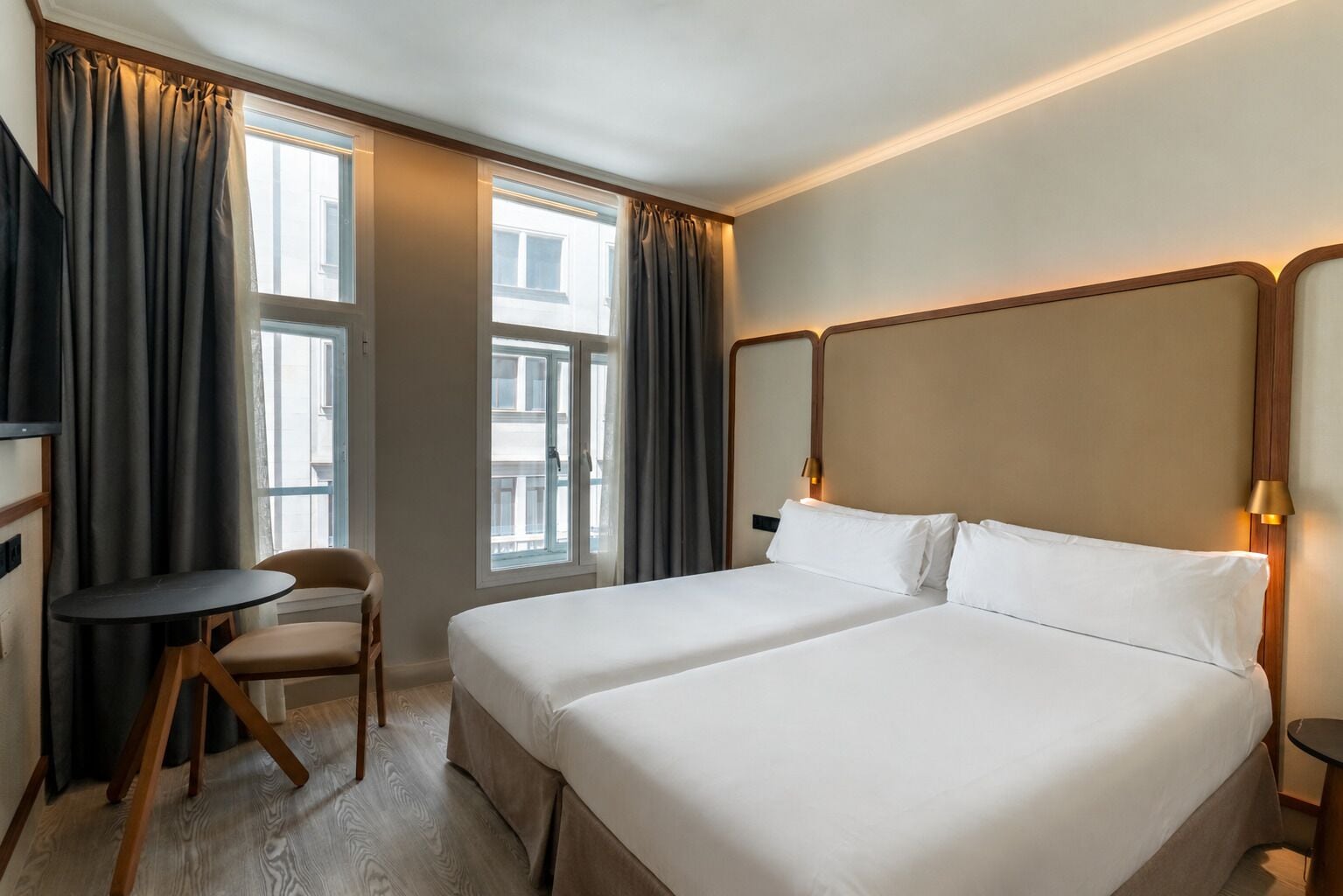 A hotel room with two beds, a small table, a chair, and a window with curtains.
