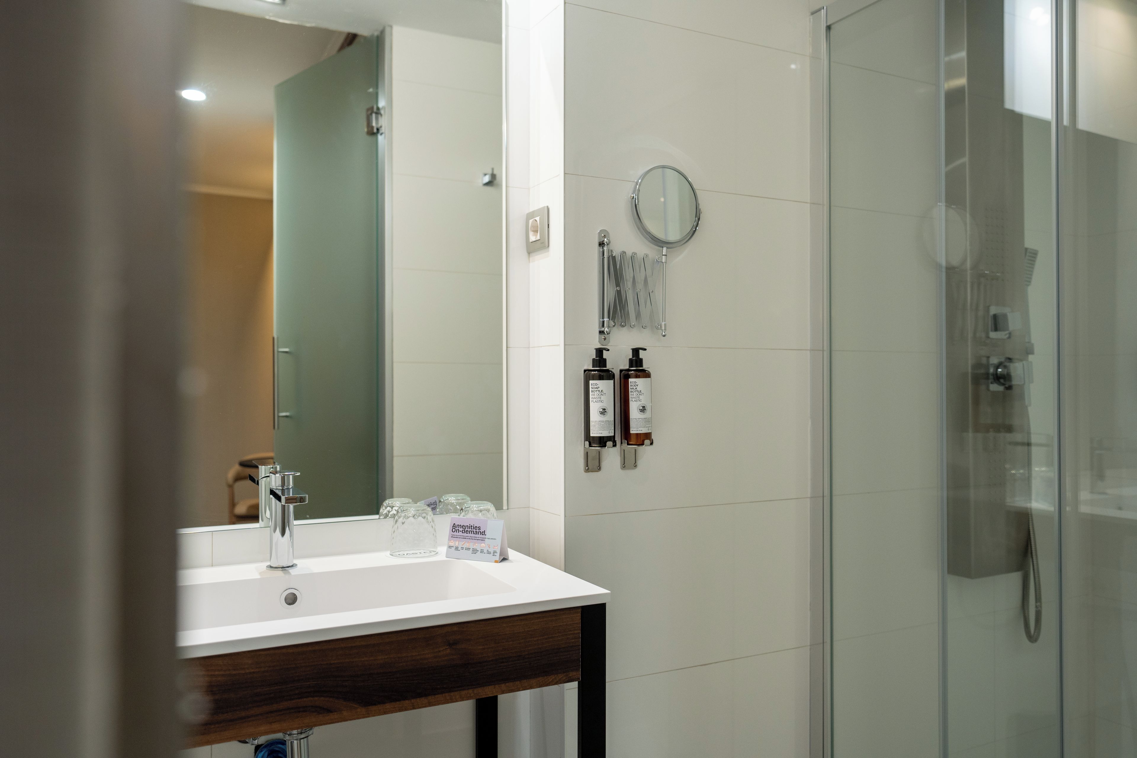 A modern bathroom with a glass shower enclosure, a white sink with a wooden countertop, and two eco-friendly soap dispensers.