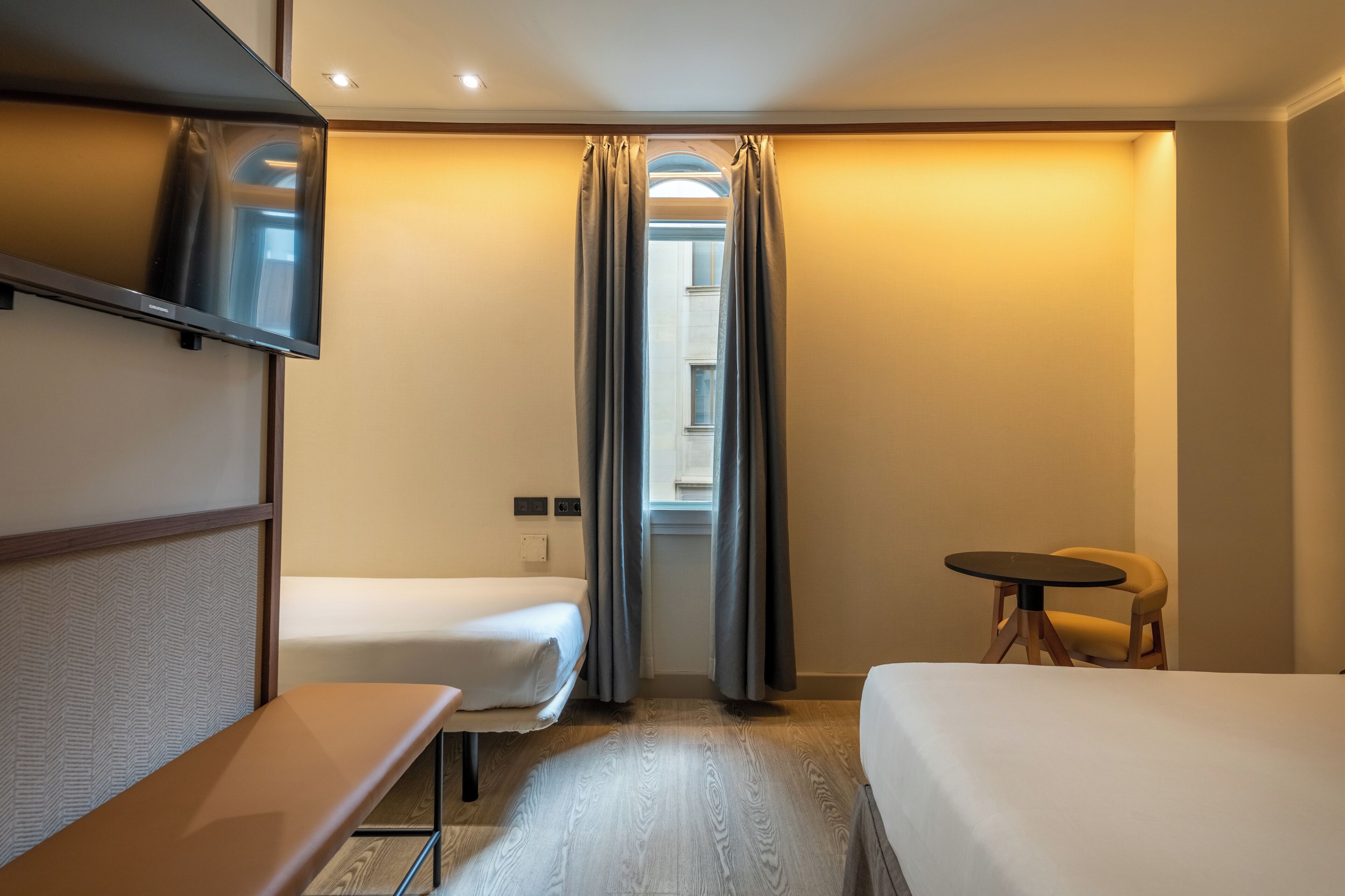 A hotel room with two beds, a small table, a chair, a TV, and a window with curtains.