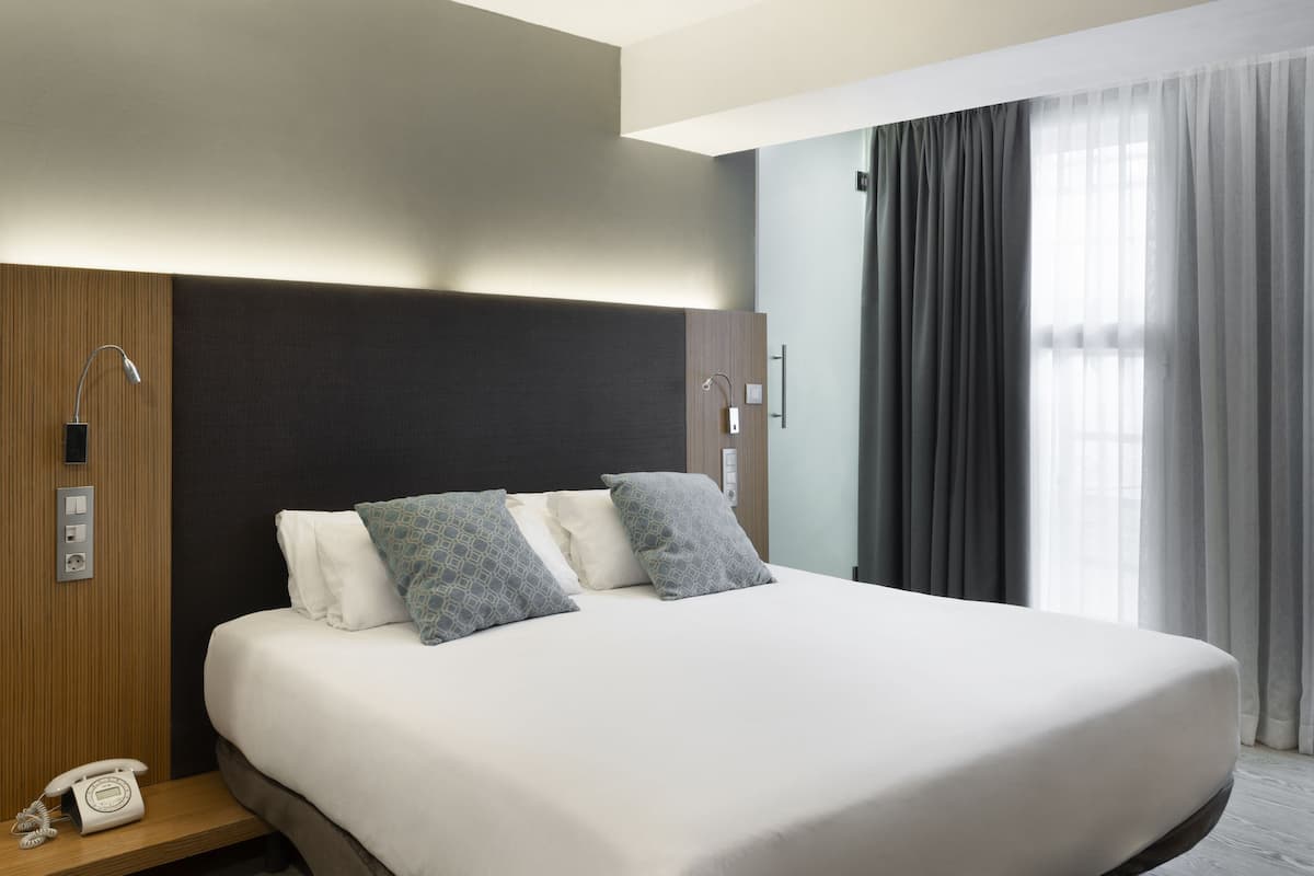 Una habitación de hotel moderna con una cama grande, un panel de pared de madera, un teléfono en la mesita de noche y una ventana con cortinas.