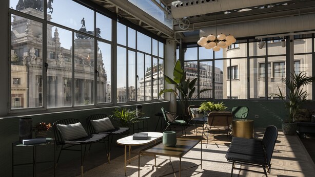 Un interior moderno con ventanas amplias, una zona de estar con sillas y mesas, y vistas a un edificio histórico.