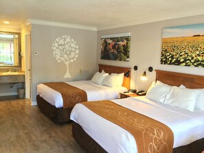 A hotel room with two beds, a large window, a tree decal on the wall, and a painting of a sunflower field.