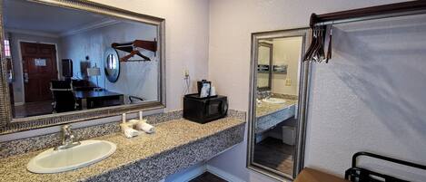 A hotel bathroom with a granite countertop, a white sink, a large mirror, a towel rack, and a bench.