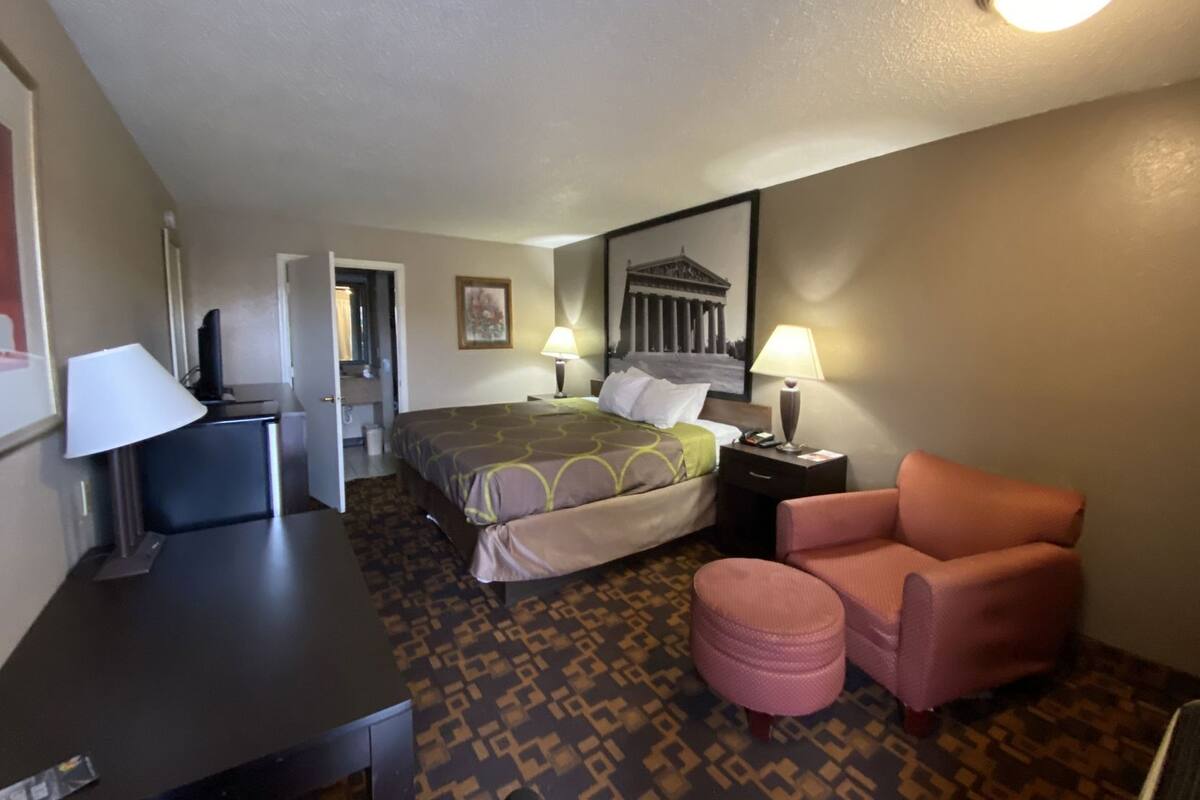 Une chambre d’hôtel comprenant un lit, un bureau, une chaise, un petit pouf et deux lampes de chevet.