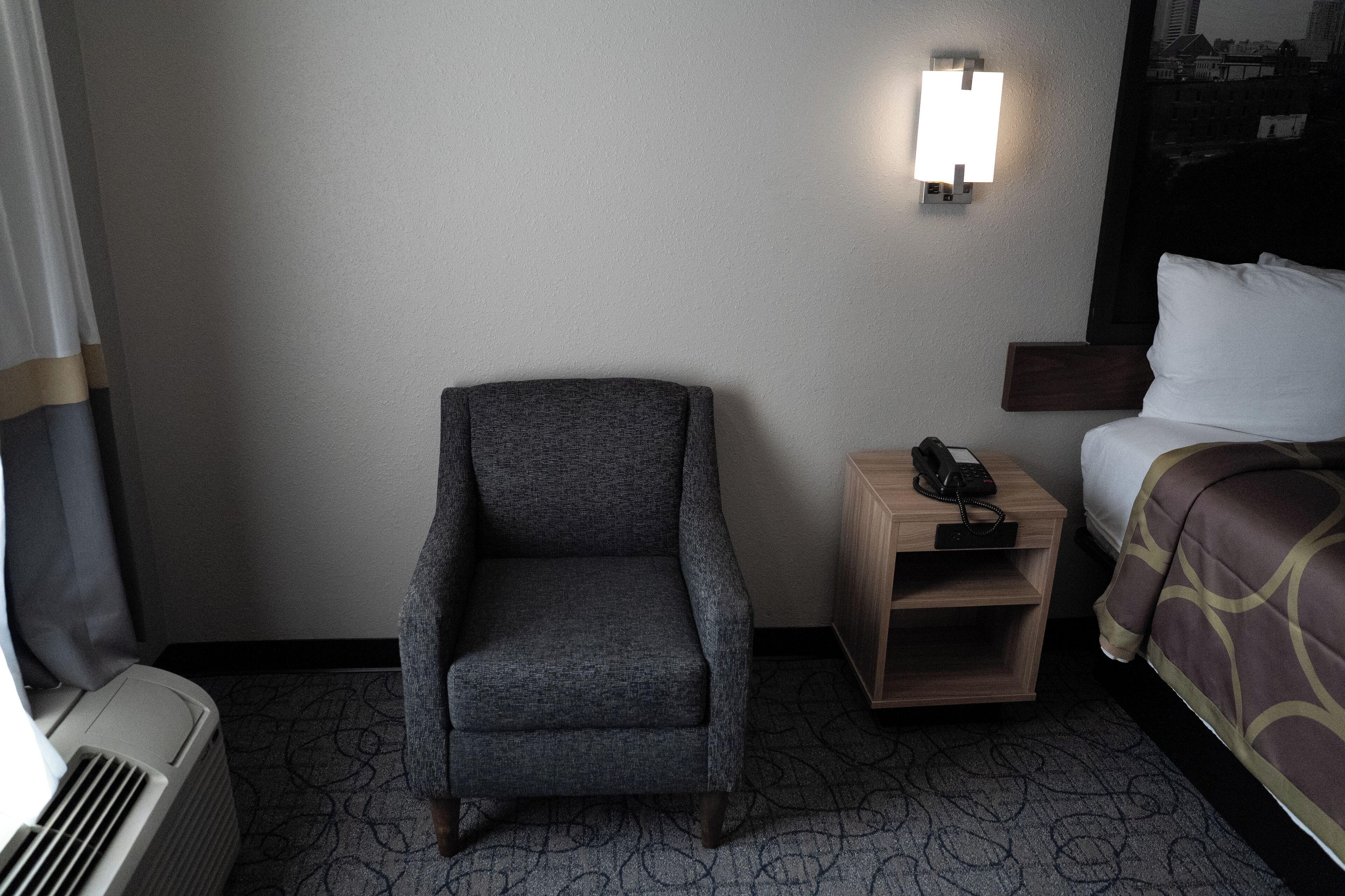 A hotel room with a gray armchair, a bedside table with a phone, a bed with a patterned bedspread, and a wall-mounted light.