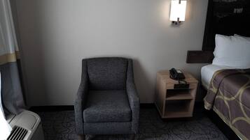 A hotel room with a gray armchair, a bedside table with a phone, a bed with a patterned bedspread, and a wall-mounted light.