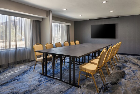 Un salón de conferencias con una larga mesa, sillas, una pantalla grande y ventanas con cortinas.