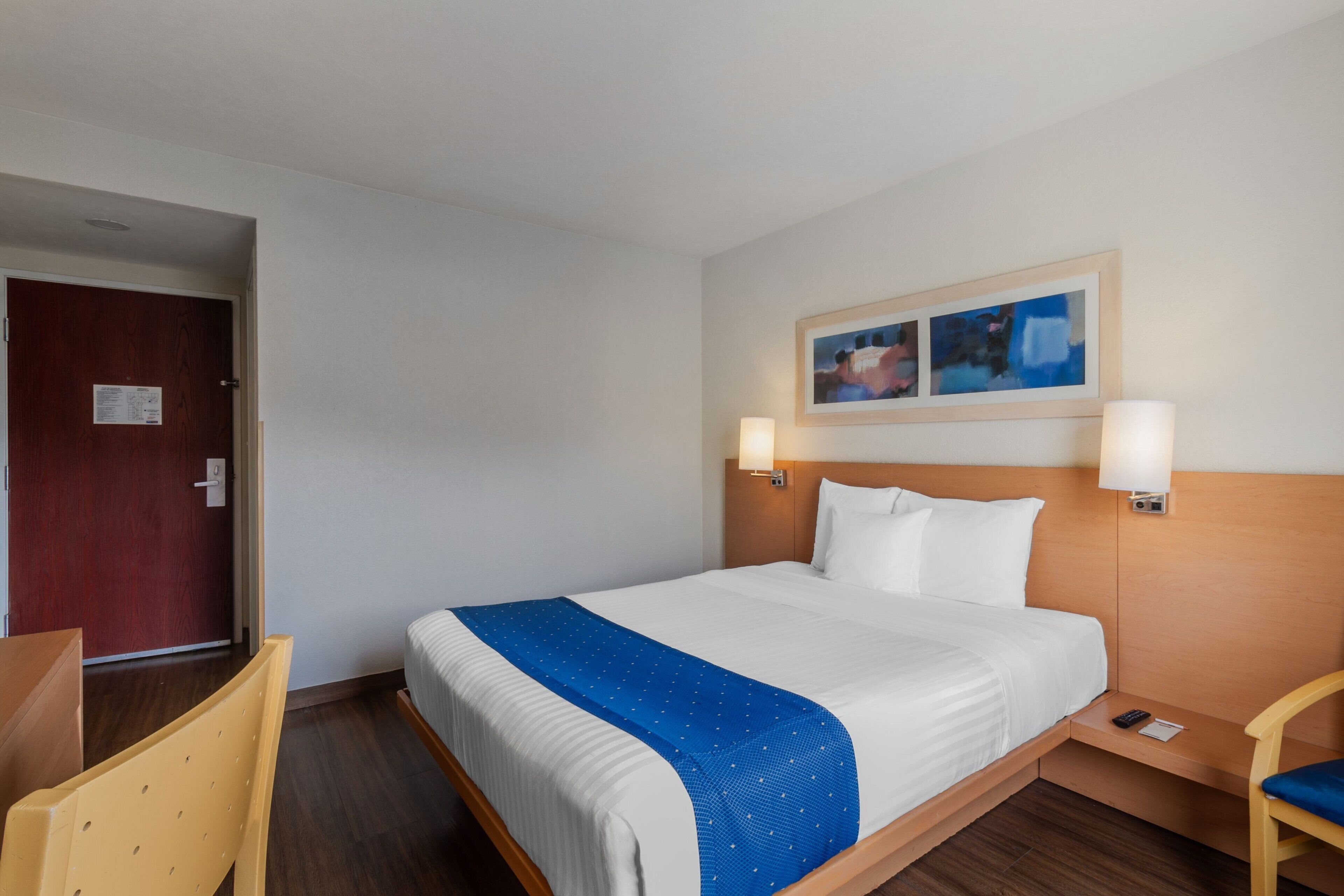 Quarto de hotel com cama, cabeceira de madeira, duas luminárias fixadas na parede, manta azul, cadeira de madeira e porta com um aviso.